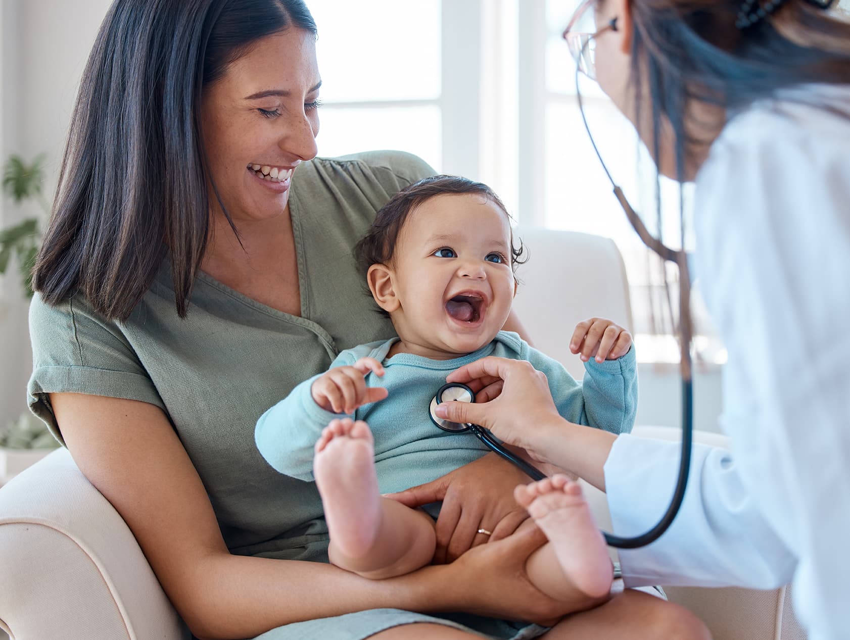 home-page-header A doctor using a stethoscope on a happy baby while the baby's mother hold's her in her lap.