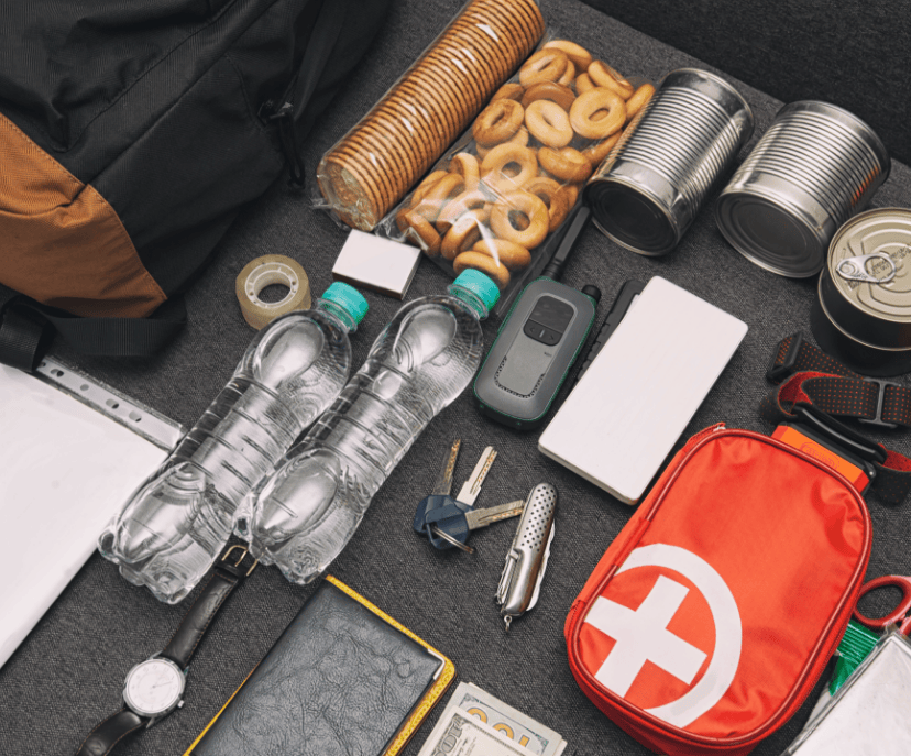 A top-down image of an emergency kit, including a first aid kit, canned foods, bottled water, keys, a pocket knife, tape, a walkie talkie, crackers, tape, and cash.