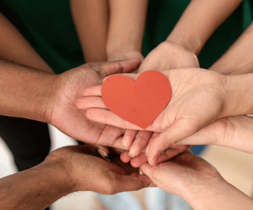 A group of hands together, palms up, holding a paper heart.